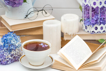 A visual for content. Still life in vintage style. A mug with a drink, an old book, a cage, candles and hydrangea flowers in the garden on a white wooden table. The concept of a tea ceremony.