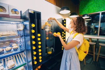 Woman in a protective medical mask squeezes orange juice using a juicer machine in a supermarket. Rules and regulation during the coronavirus pandemic