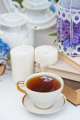 A visual for content. Still life in vintage style. A mug with a drink, an old book, a cage, candles and hydrangea flowers in the garden on a white wooden table. The concept of a tea ceremony.