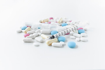 Pile of blue, white, pink and yellow colored pills and capsules with medical syringe with black markings on white background