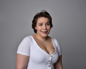 Obraz premium Close up portrait of young plus sized woman with short brunette hair, wearing a white shirt, with over the top emotional facial expressions against a light studio background. 