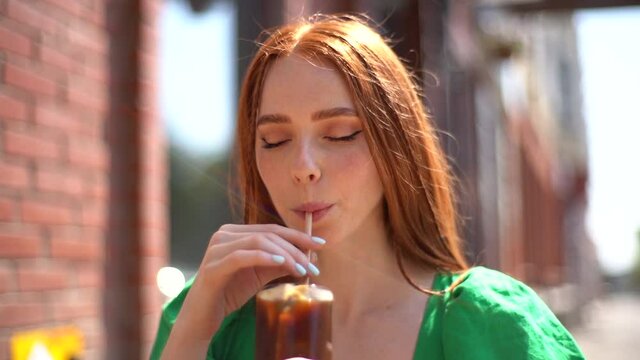 Close-up face of attractive smiling young woman drinking cocktail through straw sitting at table in outdoor cafe in sunny summer day. Headshot of beautiful happy redhead lady relaxing on urban street.