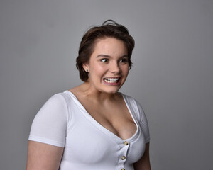 Close up portrait of young plus sized woman with short brunette hair,  wearing a white shirt, with over the top emotional facial expressions against a light studio background.  