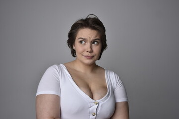 Close up portrait of young plus sized woman with short brunette hair,  wearing a white shirt, with over the top emotional facial expressions against a light studio background.  