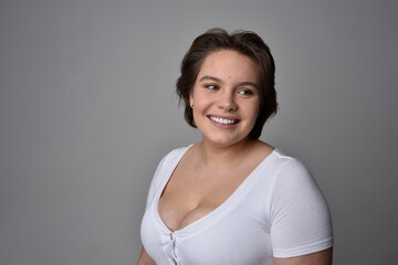 Obraz premium Close up portrait of young plus sized woman with short brunette hair, wearing a white shirt, with over the top emotional facial expressions against a light studio background. 