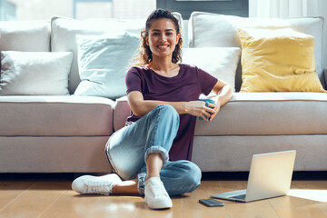 Pretty young woman using her laptop while drinking a cup of coffee sitting on the floor at home.