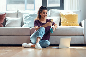 Pretty young woman using her mobile phone while working with laptop sitting on the floor at home.