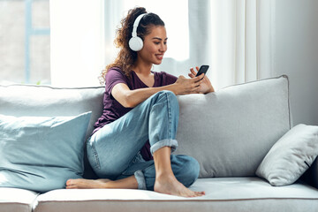 Smiling young woman listening to music with smartphone while sitting on sofa at home.