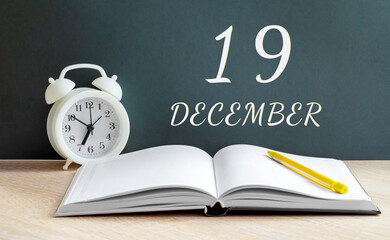 december 19. 19-th day of the month, calendar date.A white alarm clock, an open notebook with blank pages, and a yellow pencil lie on the table.Winter month, day of the year concept