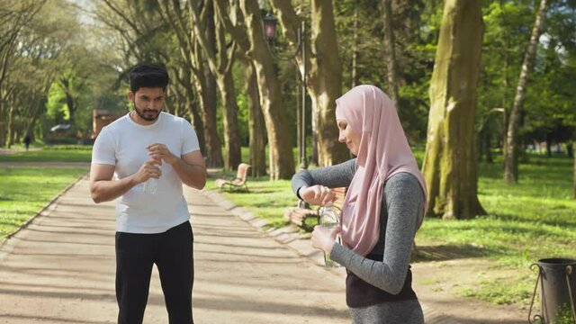 Beautiful woman in sport clothes and hijab holding bottle of water while standing at green park. Blur background of handsome muslim guy refreshing and relaxing after run. Couple in sportswear posing