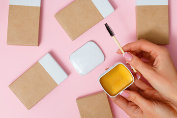 Professional cosmetics for eyebrows. Female hands hold an open jar of eyebrow soap and a brush on a pink background.