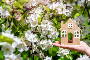 The girl holds the house symbol against the background of blossoming appletree
