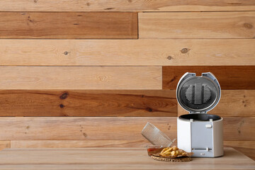 Modern deep fryer and tasty potato on table near wooden wall