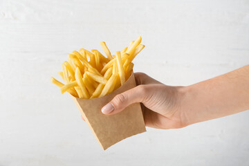 Female hand with tasty french fries in paper bag on light background - obrazy, fototapety, plakaty
