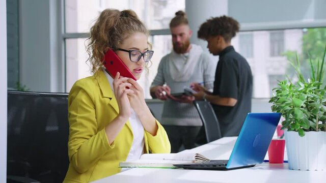 Stylish businesswoman in eyeglasses and yellow blazer talking on phone and writing down information in notebook, diverse colleagues talking on blurred background. Work day in office