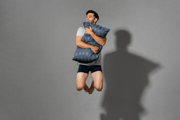 Portrait of young fresh energetic man wearing sleepwear holding pillow and jumping in mid-air after...