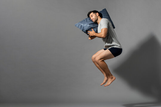 Young Sleeping Man Holding His Pillow Levitating In Empty Gray Room Background With Copy Space