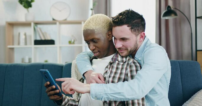 Handsome smiling happy passionate young gay embracing his black-skinned boyfriend when they together sitting on the couch at home and applying smartphone