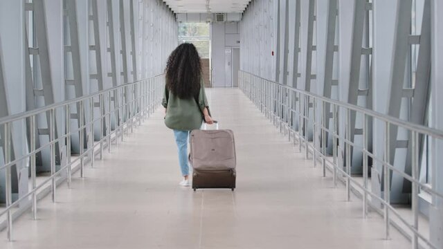 Back view unrecognizable brunette woman with long curly hair in casual clothes carries suitcase luggage walks through terminal airport steps moving to board airplane flight on journey trip vacation