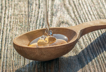 A wooden spoon in which honey is poured, while he sparkles from the rays of the sun. A spoon lies on a textured old table.
