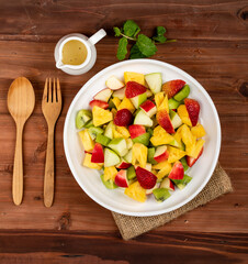 Mixed fruits salad including strawberry, kiwi, apple, and pineapple in white dish place on sackcloth on wooded table.  Utensil and oil salad dressing cup beside with mint leaf decorating