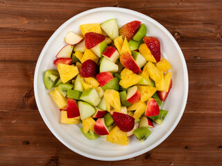 Mixed fruits salad including strawberry, kiwi, apple, and pineapple in white dish place on dark clean wooded table. vegetarian dish