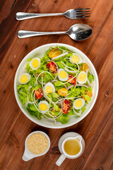 Big white bowl of lettuce, sprout salad with boil egg, onion and tomato mix with Japanese sesame oil dressing on wooden table together with small olive oil jar and sesame cup and utensil beside