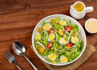 Big white bowl of lettuce, sprout salad with boil egg, onion and tomato mix with Japanese sesame oil dressing on wooden table together with small olive oil jar and sesame cup and utensil beside