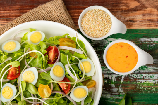 Cloth up picture of big white bowl of lettuce, sprout salad with boil egg, and tomato mix with sesame dressing on sack cloth above wooden table and small olive oil jar and sesame cup