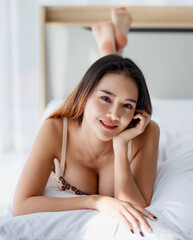 Obraz na płótnie Canvas Beautiful young naughty Asian girl with sexy bra enjoy lie down with pillow on white bed and impassioned smile for bright morning of rest day.