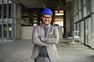 Smiling businessman with helmet on head standing with arms crossed in his building in construction process and looking at camera.