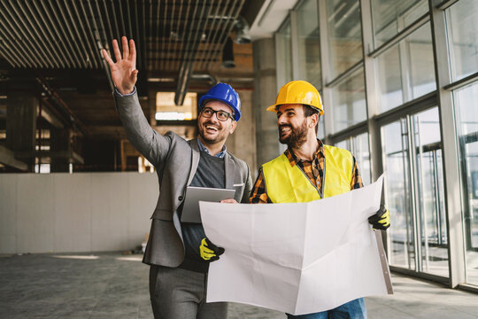 Smiling architect holding tablet and showing to construction worker something he imagined. Construction worker holding blueprints. Construction site interior.
