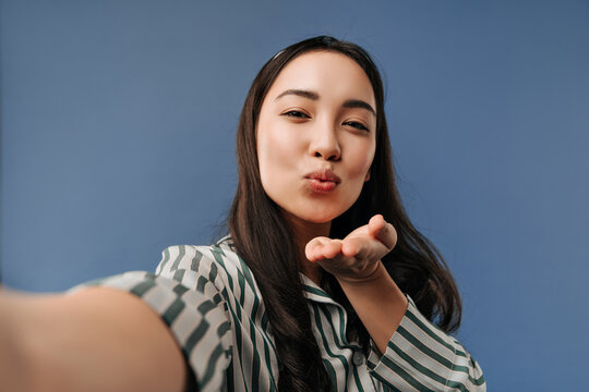 Stylish cool asian girl long dark hairstyle in fashionable white and green pajamas blowing kiss and taking photo on blue backdrop..