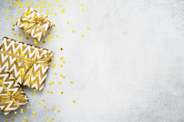 Golden Christmas gifts and star shaped confetti on grey background. Birthday or anniversary present box in paper with geometric print and shiny decorations flat lay. New Year composition. Copy space.