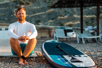 Tanned handsome man sitting at the pebble beach with a sup board. Wooden umbrellas and chaise longues at defocused background. Surfing and activity