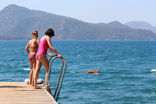Beach holiday on sea resort, kid girls in swimwear going to jump into the water from wooden pier. Scenic view to the green mountain coast in mist