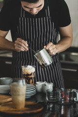 Fototapeta na wymiar Young Asian barista man on black-and-white apron using spoon to deliberately topping coffee with milk froth for softness and mellowness at counter surrounding by dishes, jars, glass