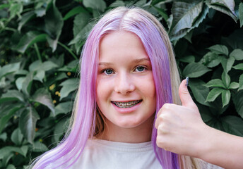 Smiling teen girl wearing dental braces with her thumb up, cute caucasian girl with pink hair...