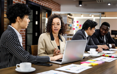 Close up to two mixed race caucasian african beautiful smart professional businesswomen talking, discussing job strategy, using laptop for working in indoor office with background of male colleagues.