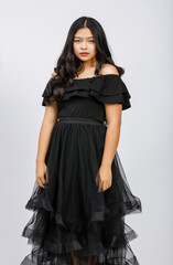 Vertical portrait shot of a cute smiling young girl teenager in a black dress looking at camera in studio isolated white background. Junior girl in fashionable clothes touching hair with shy emotion