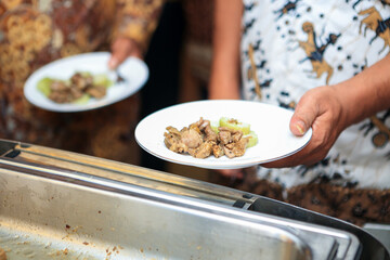 Wedding party guest queue at food stall holding white plate containing lamb chops and rice cake with bokeh background. Selective focus.