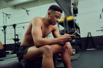 Fototapeta na wymiar Young African American male looking at his phone to reply to an online message. Mixed race, male personal trainer looking at his phone while sitting down in the gym. High quality photo 