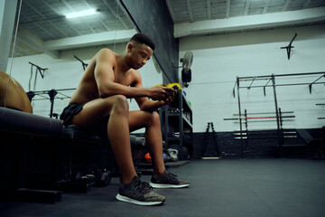 Obraz premium Young African American male looking at his phone to reply to an online message. Mixed race, male personal trainer looking at his phone while sitting down in the gym. High quality photo 