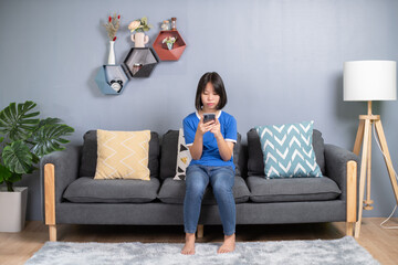 asian girl sit on couch in living room holding smart phone, chatting use social media network, watch received video from friend, with modern wireless technology.