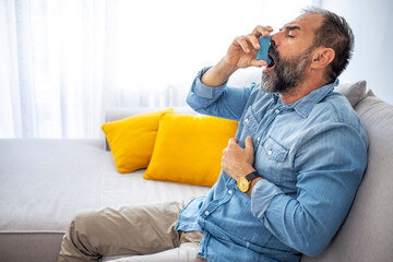 Side view of an asthmatic man using an inhaler sitting on a couch in the living room at home. Man...