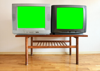Two antiquated vintage TVs with blank green screens sit on a vintage table in a 1990s tenement building.