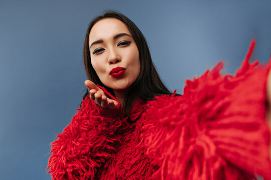 Trendy charming girl with long brunette hairstyle and bright makeup in fluffy warm red sweater blowing kiss and taking photo..