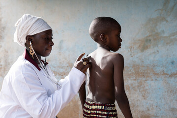 Attentive black doctor placing a stethoscope on the bare back of a standing thin schoolboy during a...