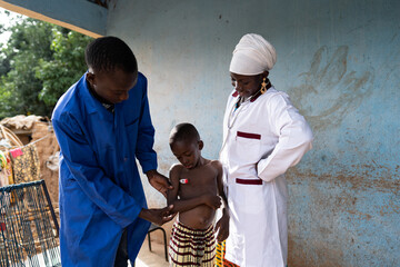 Medical trainee in blue workdress taking a small toddler's temperature under the close supervision...