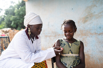 Attentive young African nurse in a white coat holding a digital thermometer under the armpit of a...